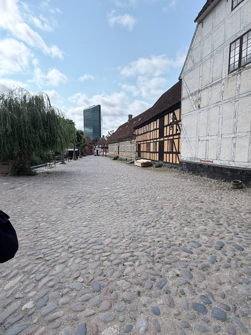 Cobblestone lane in Den Gamle By with a weeping willow tree and modern tower in background