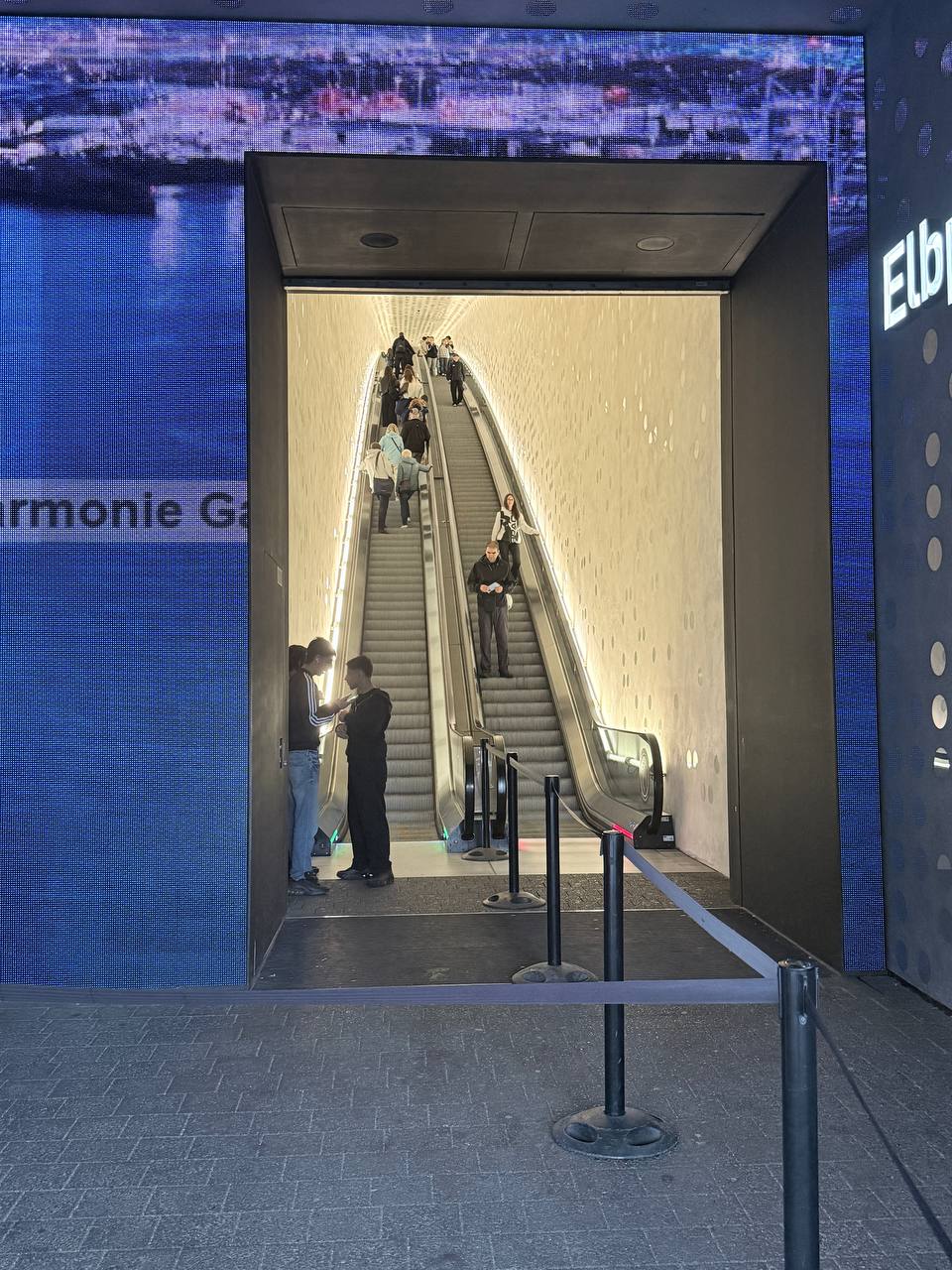 Escalator tunnel entrance with digital screens showing cityscape and Elbphilharmonie imagery