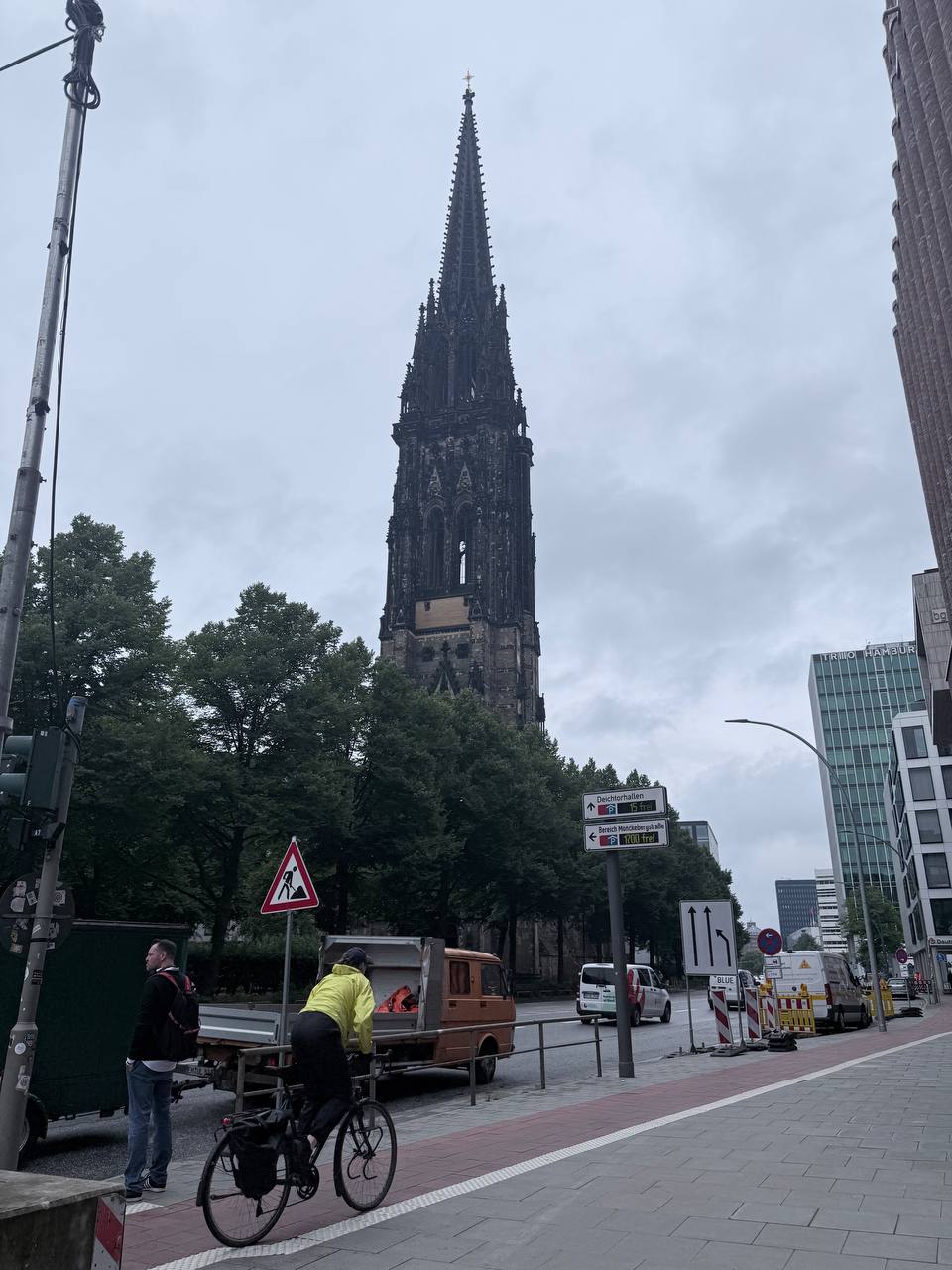 Gothic church spire with cyclist on red brick sidewalk surrounded by cranes and modern buildings