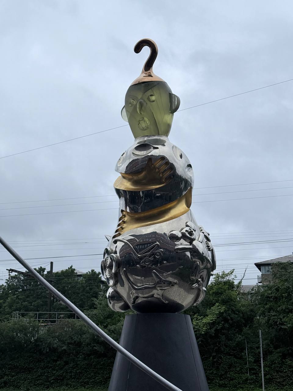 Abstract metallic sculpture with translucent olive-green head, polished silver body with golden band, and reflective chrome base featuring sculptural animals and gears