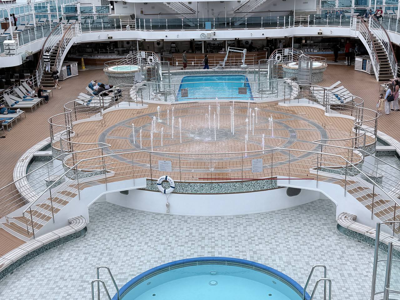 Multi-level cruise ship pool deck with central fountain, two hot tubs, swimming pool and sun loungers