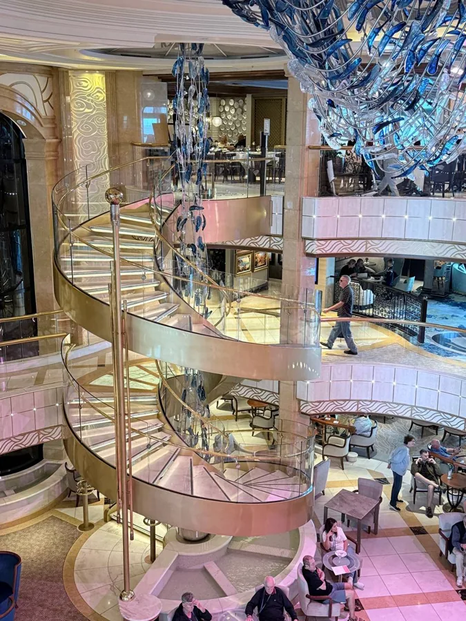 The Majestic Princess atrium - a grand double-helix spiral staircase with gold handrails, fish-shaped chandeliers, marble floors, and a water feature at the base