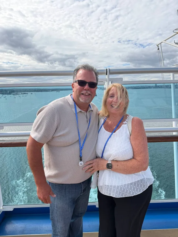 Couple smiling on cruise ship deck at sail away with waterfront and wake behind them