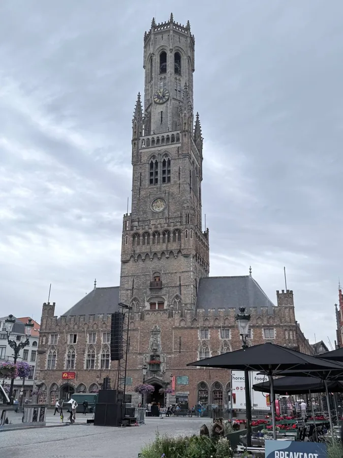 The 83-metre Belfry of Bruges rising above the Grote Markt, with Gothic windows, clock faces, and a sky of fluffy white clouds