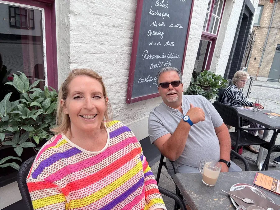 Two holidaymakers enjoying coffee at an outdoor café, giving double thumbs up, with a chalkboard menu on a whitewashed wall behind them