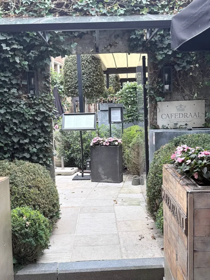 The ivy-covered arched entrance to Cafedraal restaurant in Bruges, with stone-paved path, climbing ivy, and pink flowers