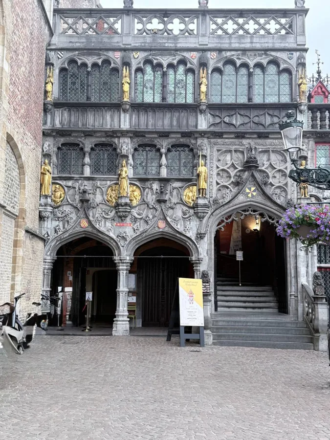 Ornate Neo-Gothic building facade with golden statues in niches, diamond-paned windows, carved stonework, and a cobblestone foreground