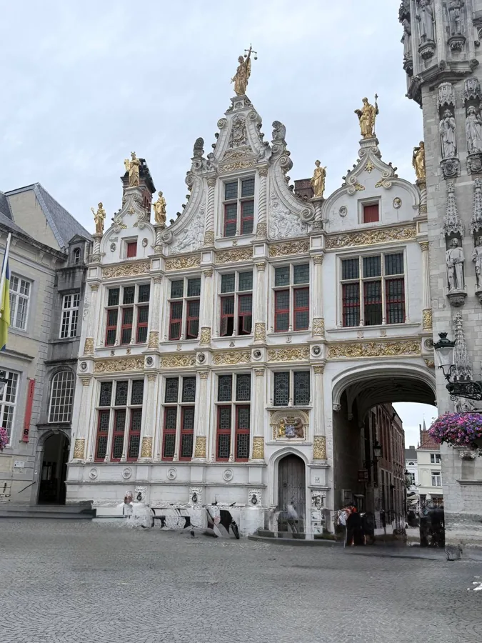 Grand Baroque-style building with white and gold facade, three golden statues on the roofline, red-framed windows, and a cobblestone square