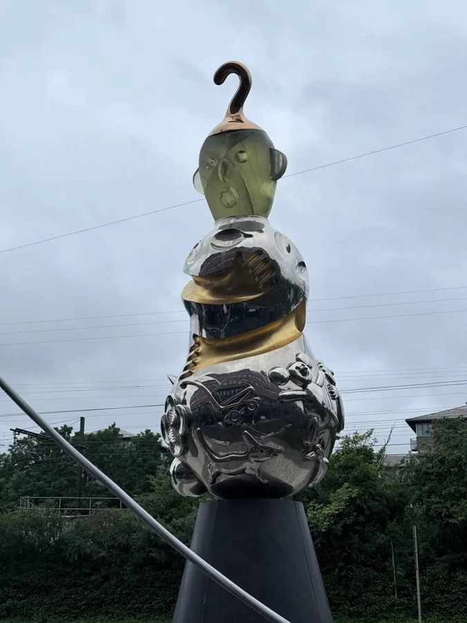 Abstract metallic sculpture with translucent olive-green head, polished silver body with golden band, and reflective chrome base featuring sculptural animals and gears