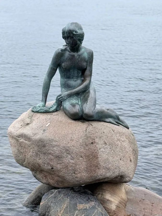 The iconic Little Mermaid statue in Copenhagen, bronze with greenish patina, sitting gracefully on a granite rock with calm harbour water behind her