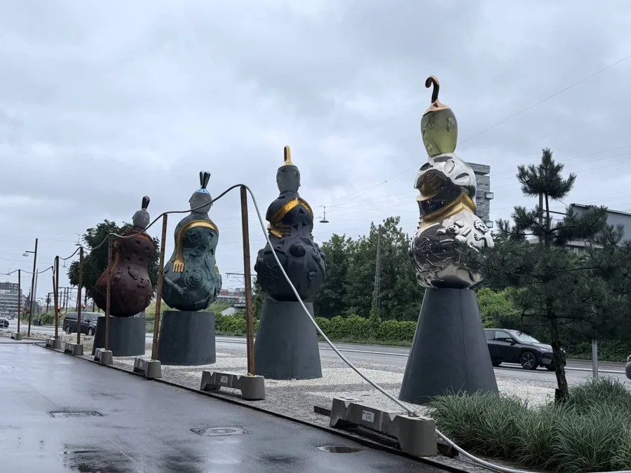 Four abstract modern sculptures in a row on a wet sidewalk, showing an evolution from earthy red-brown patina to polished silver, connected by white cables
