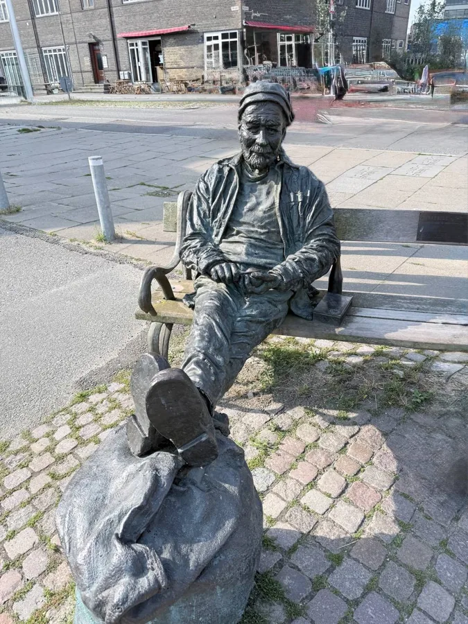Bronze statue of an elderly man seated on a wooden bench on Aarhus harbour front, with cobblestones and buildings in background