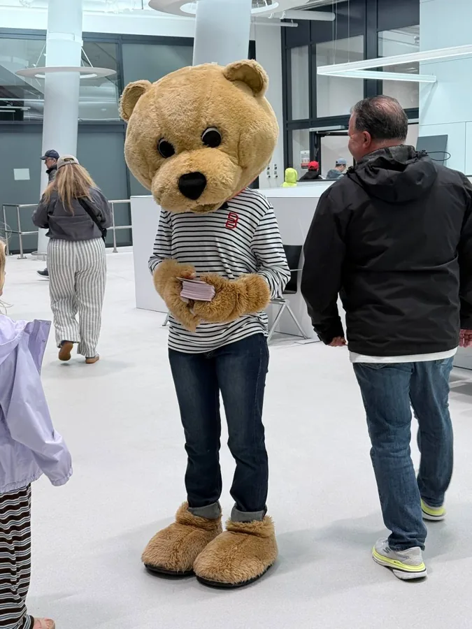 Person in a teddy bear mascot costume holding cards in a modern lobby